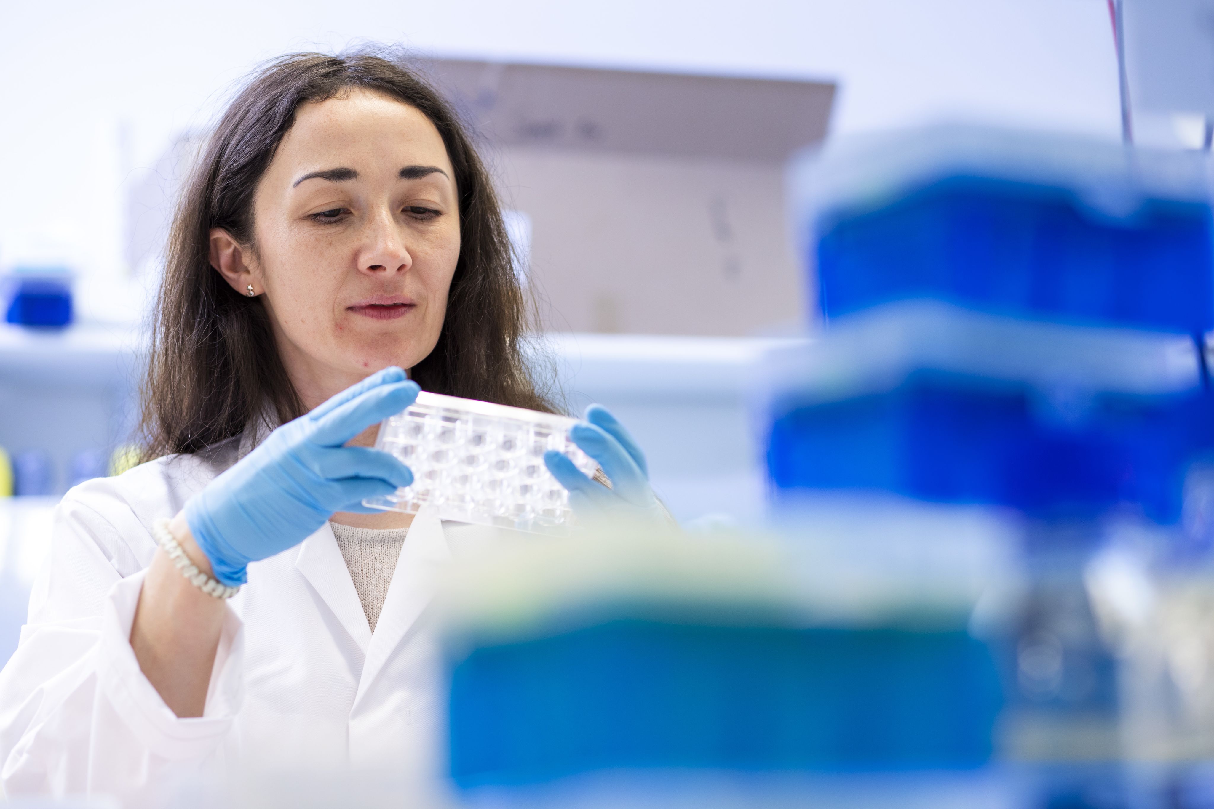 A young woman with dark hair, wearing blue sterile gloves and a lab coat examining a clear tray.
