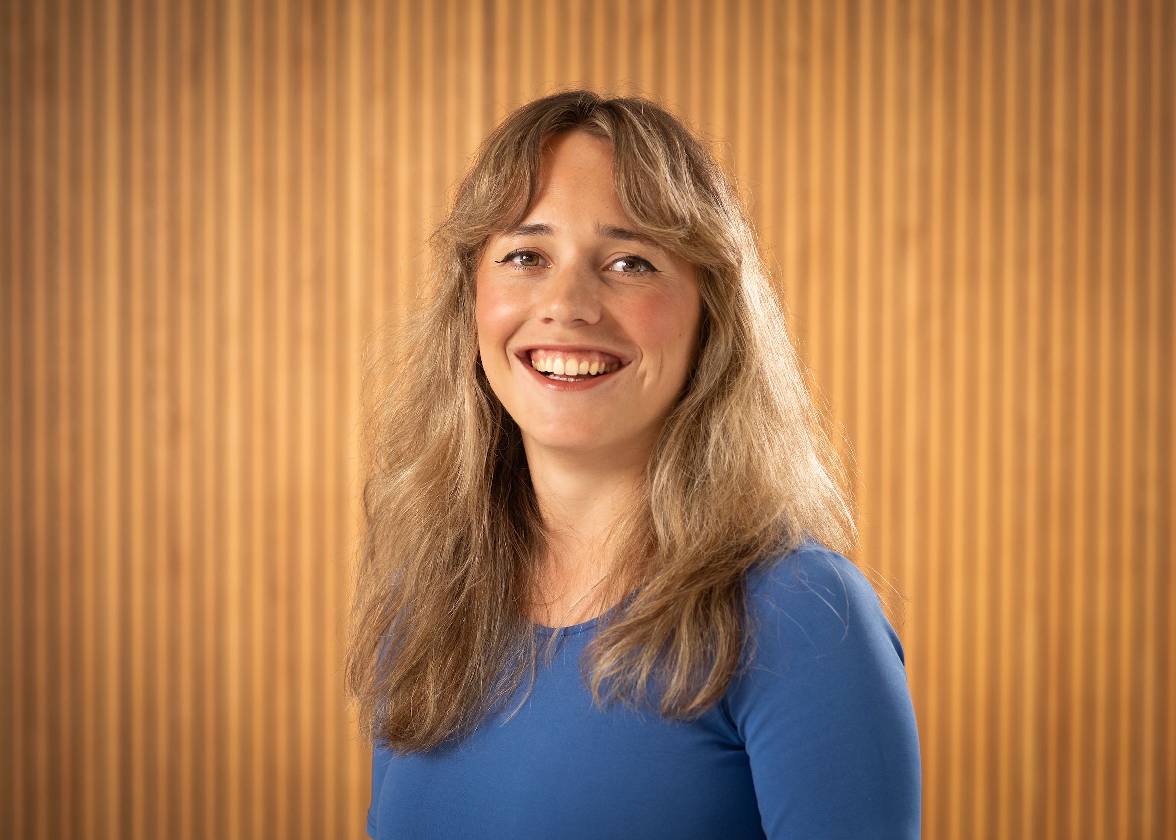 A young woman blonde hair and a blue shirt facing the camera directly and smiling.