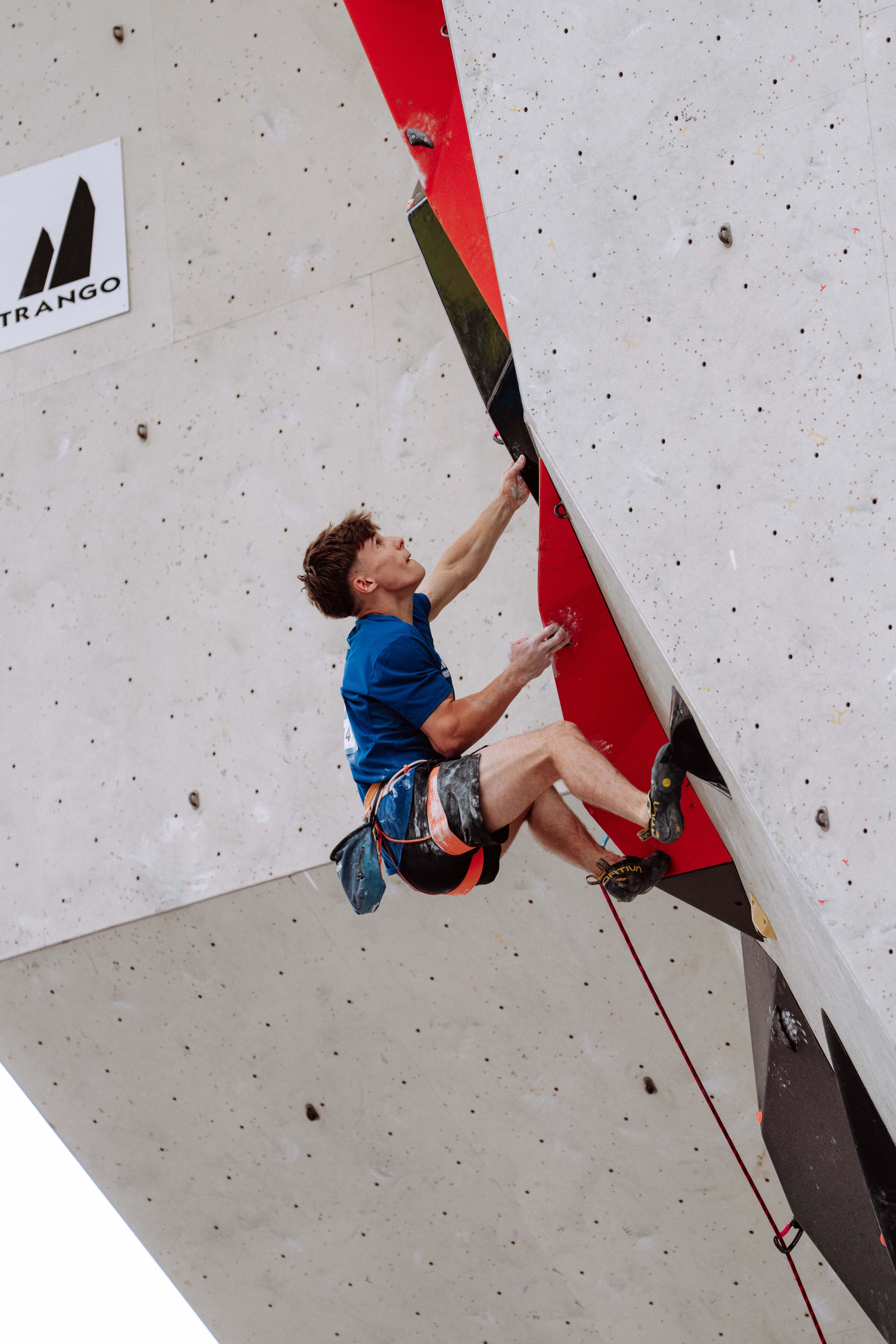 A young man with a serious expression, wearing a blue shirt and a harness climbing an indoor rock climbing wall
