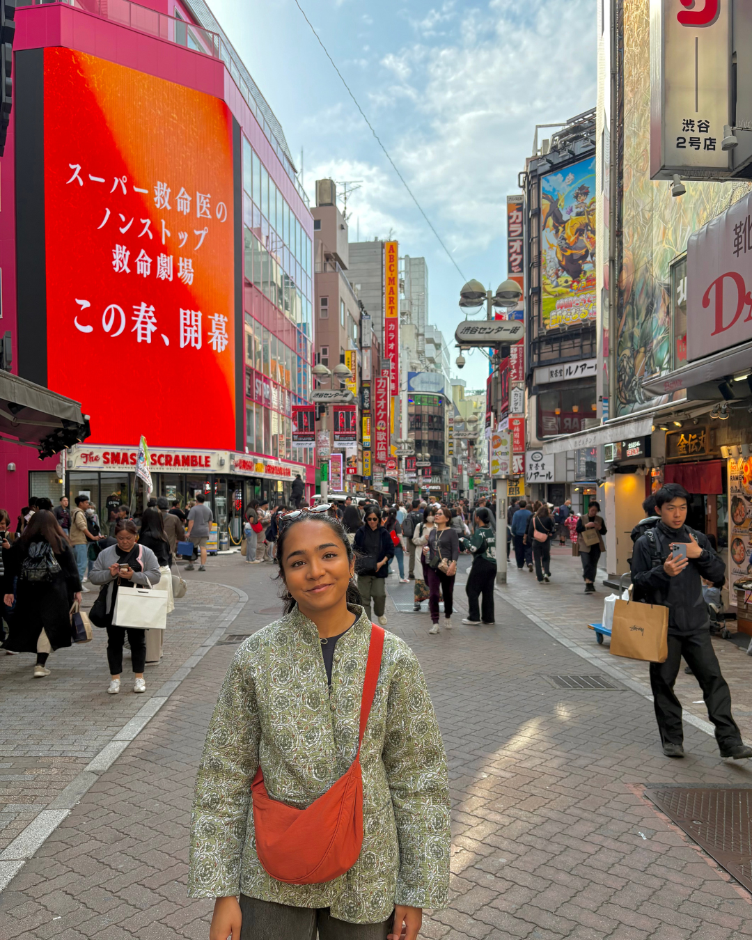A young woman in a green jacket, wearing an orange sling bag stands outside in a busy alleyway in Japan