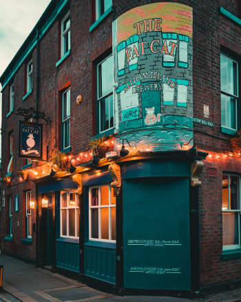 A pub with glass windows and doors, a board with text on the wall, lights, plants, and other objects. At the bottom of the image, there is a road, and in the background, the sky can be seen. The building is the Fat Cat Pub in Kelham Island, Sheffield.