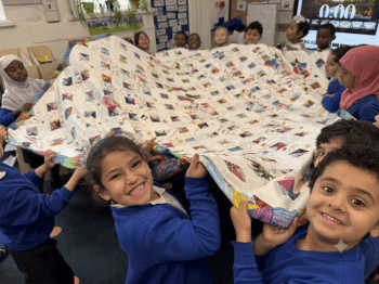 A group of children holding up a large quilt in a room with a wall in the background, a window to the left, a plant to the side, and a screen to the right.