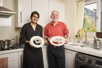 A blind man and female student standing in a kitchen, both of them holding plates in their hands. The kitchen is equipped with a washing machine, a gas stove, utensils, a sink with a tap, a bottle, a basket, a cloth, a window with a curtain, and cupboards. Through the window, we can see trees, buildings, and the sky.