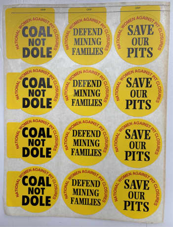 A number of yellow stickers with a white background and a bunch of yellow stickers on it. The text reads "Coal Not Dole," "Defend Mining Families" and "Save our Pits."