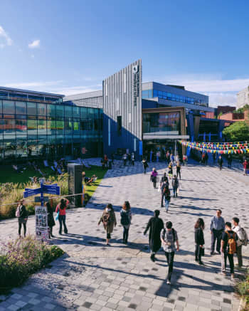 A group of students walking on the concourse in front of the SU building with glass walls. Some of the people are wearing backpacks and carrying handbags, while others are sitting on chairs. There are also plants and trees in the background, as well as decorative flags with ribbons. The sky is filled with clouds.