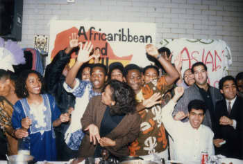 A group of people standing around a table with plates of food, glasses, bottles, and other objects. In the background, there is a banner with text on it, a speaker, clothes, and a wall.